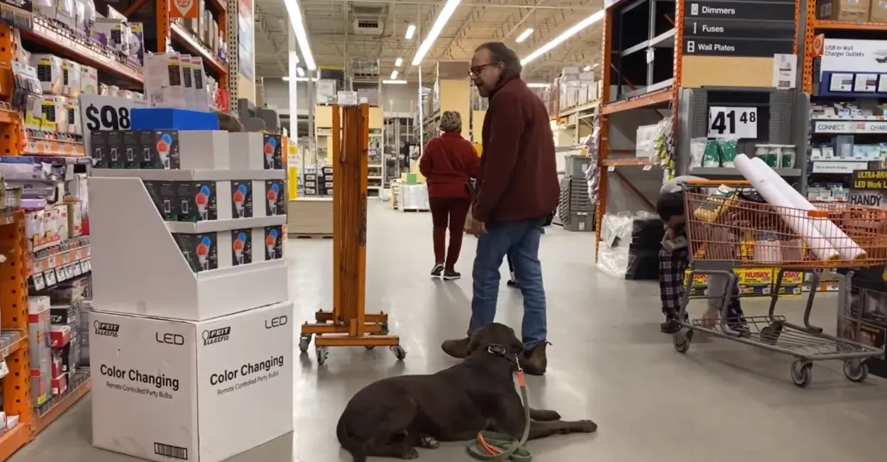 A man with a dog on a leash, trained through dog training testimonials, pauses in a hardware store aisle, looking at shelves with a woman and shopping cart visible in the background.